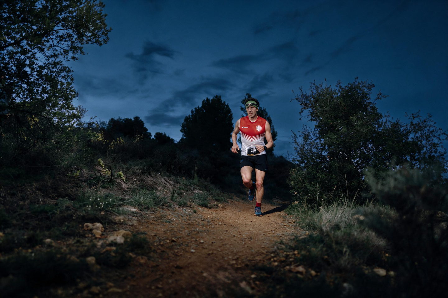 A runner with a lit headlamp on a pine-forest trail at dusk